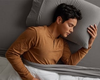 A man falls asleep fast on his side on a bed with gray sheets and white duvet.