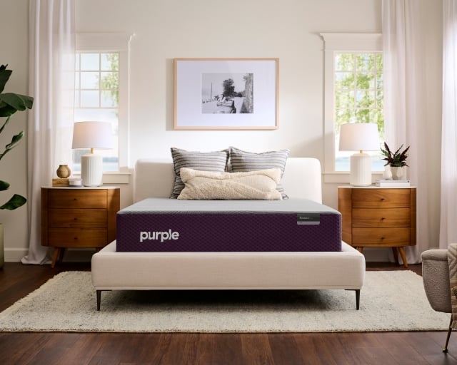 Unmade Purple Restore Plus mattress on a cream upholstered bed frame between two wood nightstands.