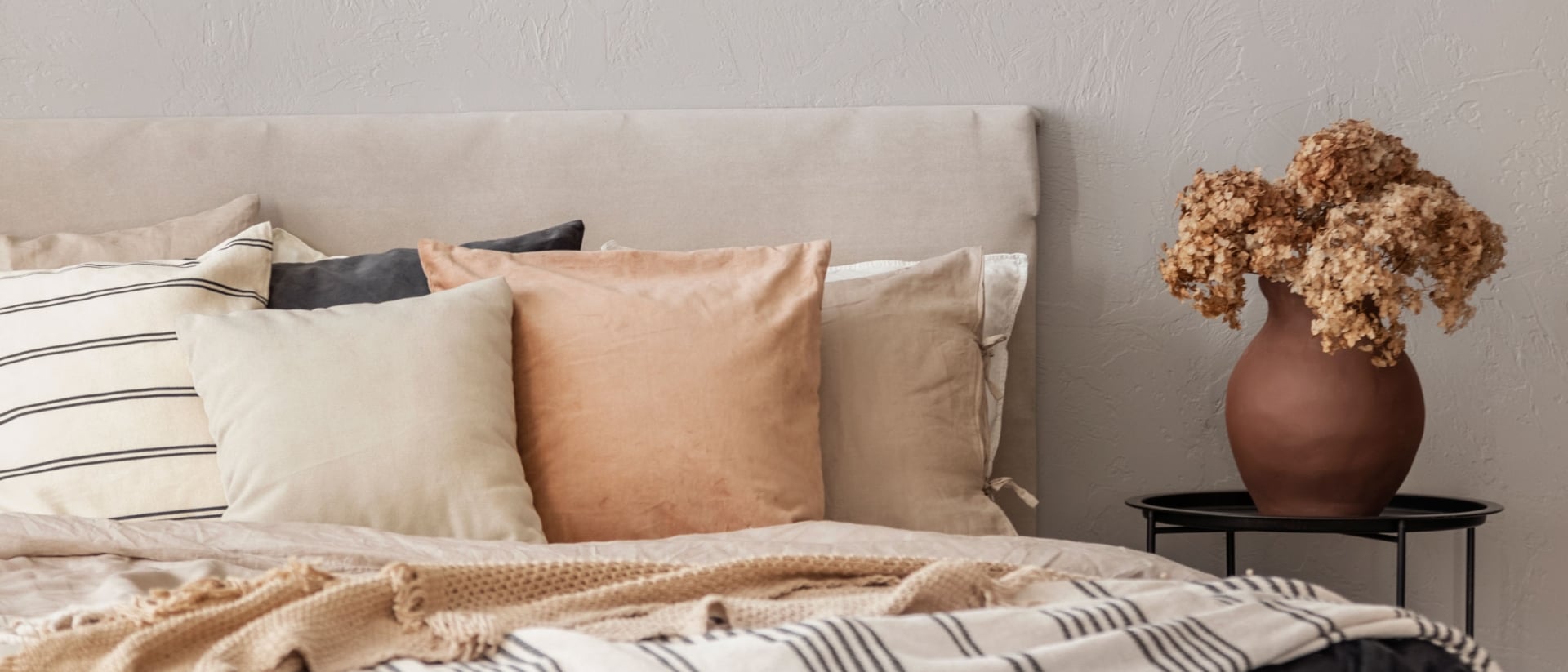 A cozy bed with neutral-colored throw pillows and blankets next to a vase of flowers on a bedside table.