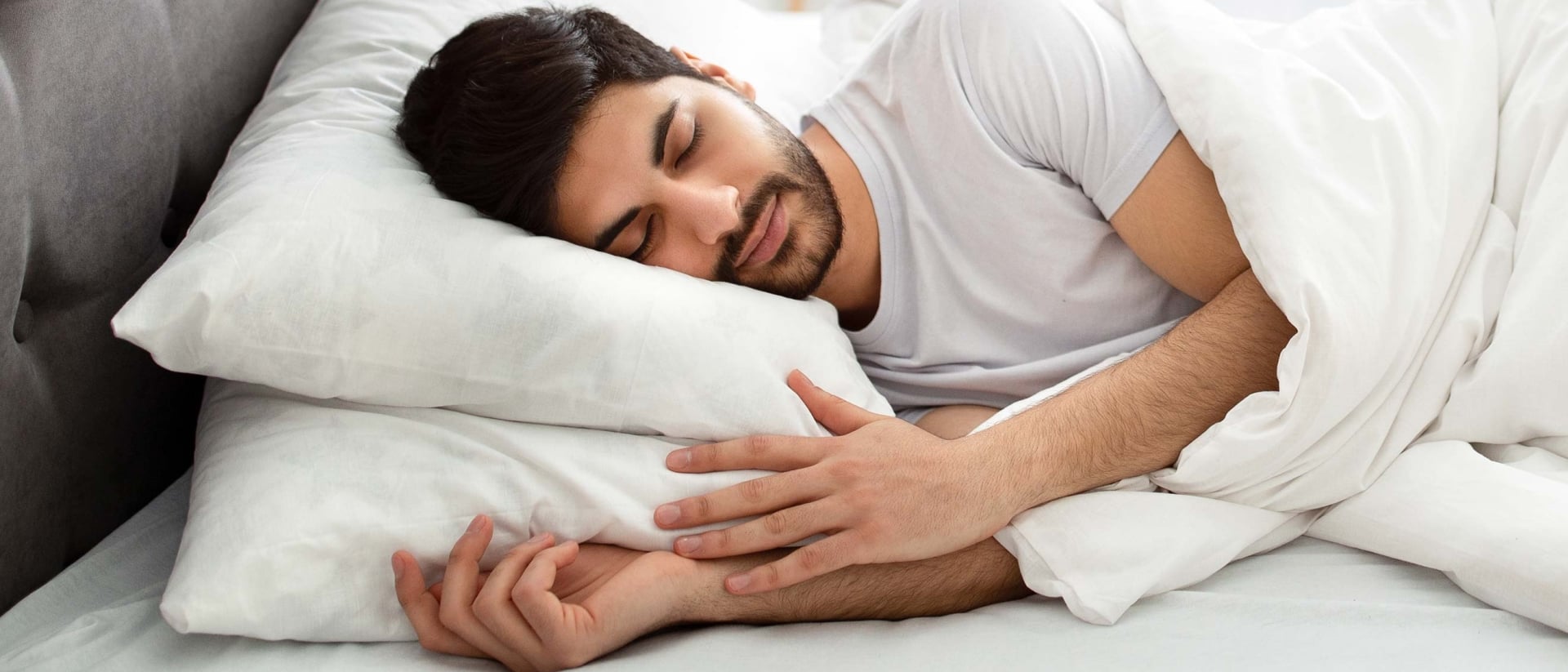 A man sleeping on his side cover with two pillows and covered in a white duvet.